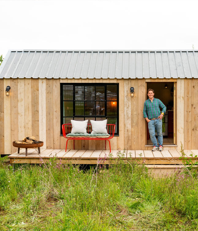 A modern wooden cabin with a metal roof features a cozy porch, a red bench with cushions, and a tranquil green landscape.