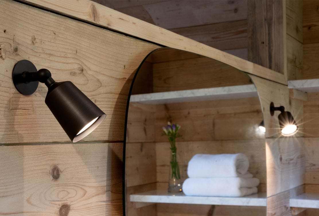 A stylish bathroom corner featuring a sleek black wall light, wooden walls, and neatly stacked white towels with a vase of flowers.