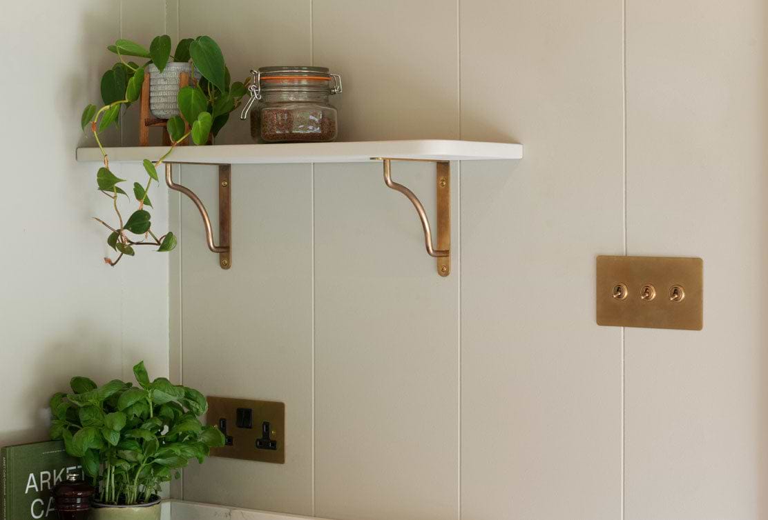 A minimalist shelf with brass brackets holds a jar of spices beside a potted plant against a neutral wall with light switches.
