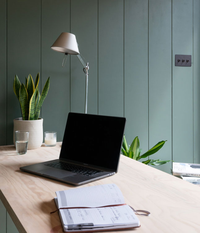 A minimalist workspace featuring a laptop, a notebook, glasses of water, and a potted plant, with green walls in the background.