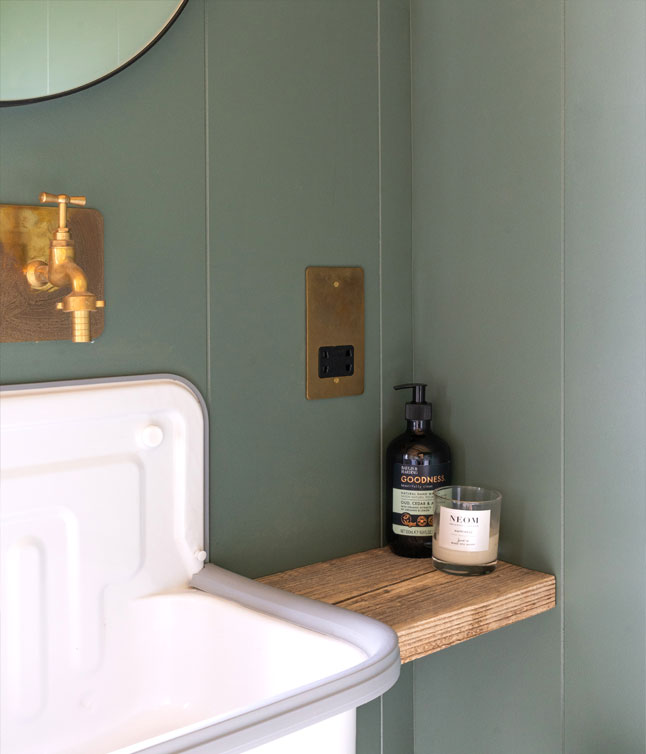 A stylish bathroom corner featuring a round mirror, wall-mounted faucet, wooden shelf with a lotion bottle and candle, against green walls.