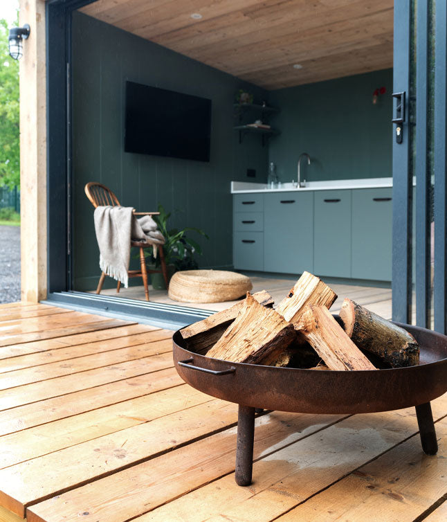 Cozy outdoor scene featuring a fire pit with logs, wooden deck, and a modern interior with kitchen and seating area visible through open doors.