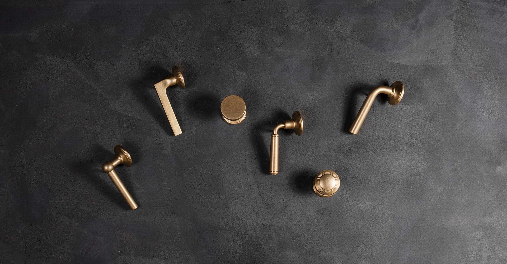 A flat lay of various brass door handles arranged artistically on a textured dark surface, showcasing different designs and shapes.