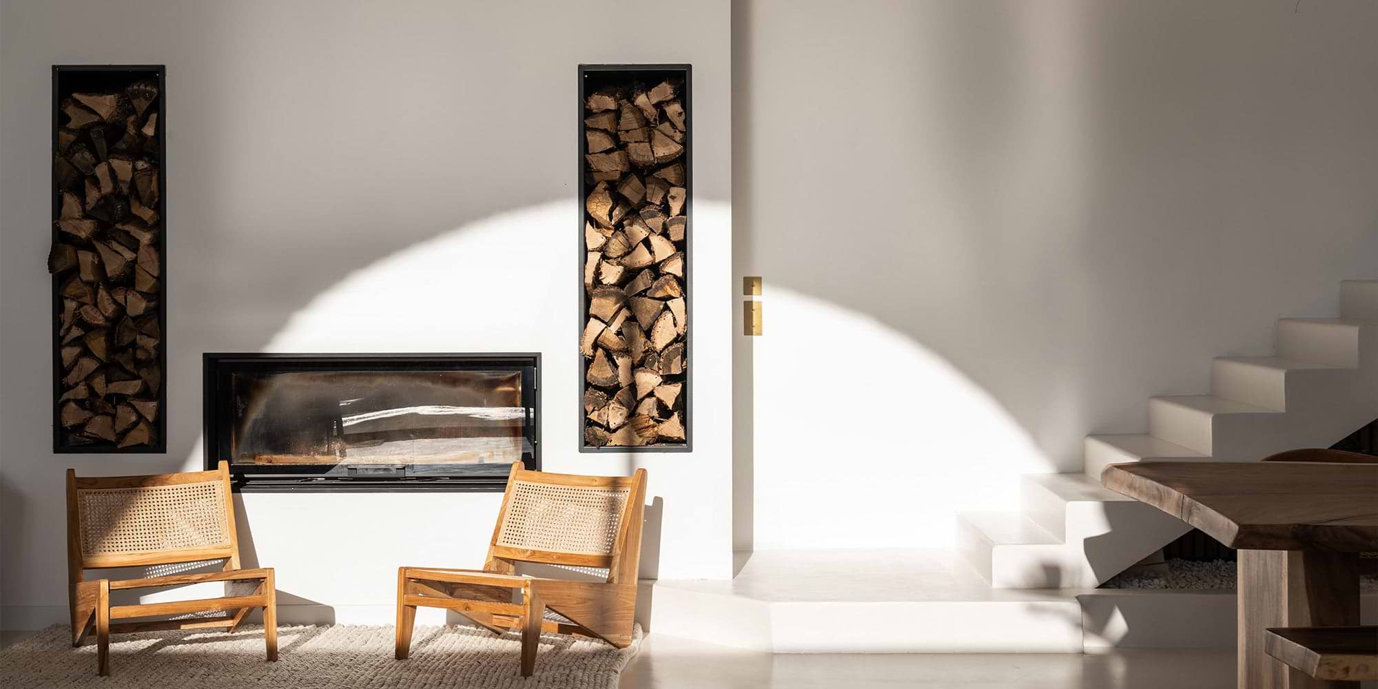 A minimalist living space featuring wood accents, two wooden chairs, a fireplace, and stacked firewood, illuminated by soft sunlight.