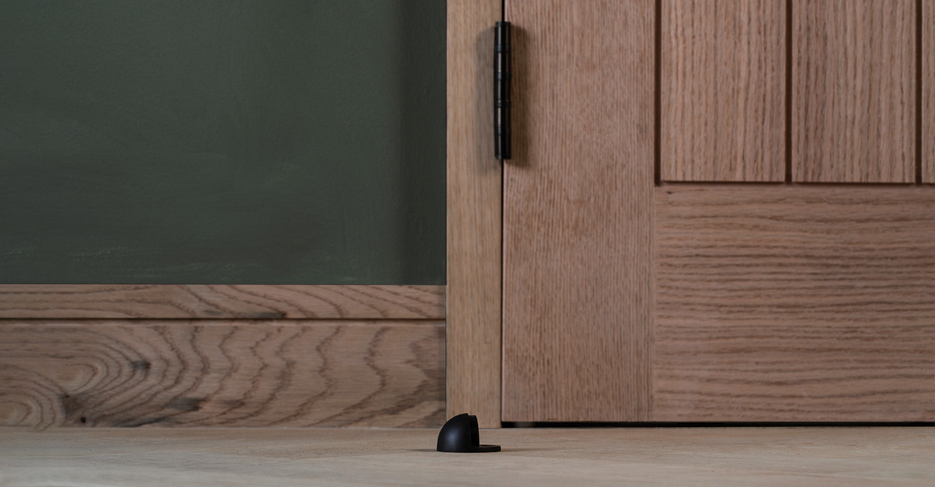 A close-up of a black door stop positioned on a light wooden floor, next to a large oak door against a dark green wall.
