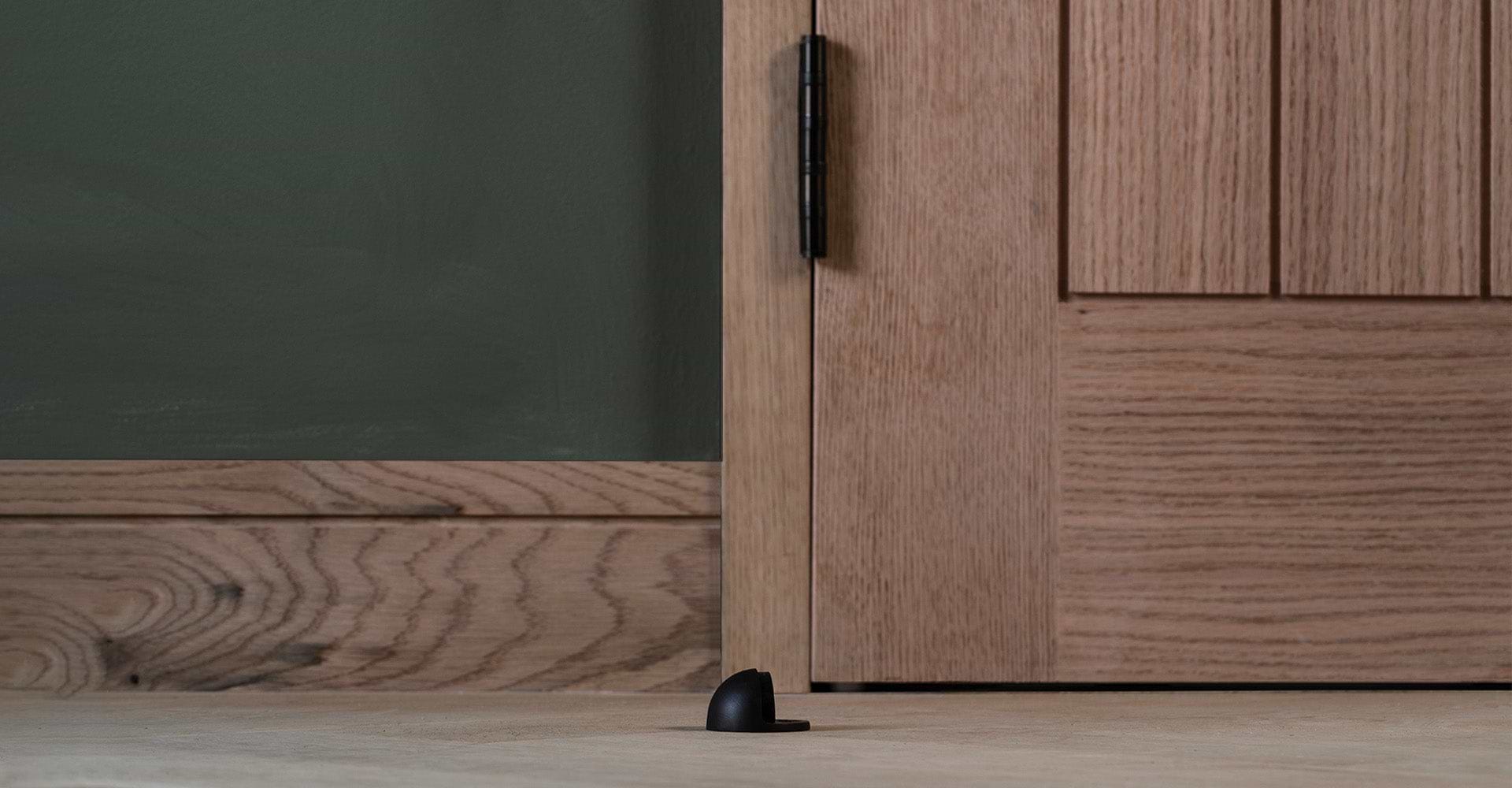 A close-up of a black door stop positioned on a light wooden floor, next to a large oak door against a dark green wall.