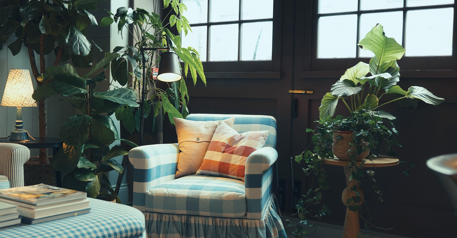 A cozy blue checkered armchair with colorful pillows, surrounded by lush plants and soft lighting, creates a tranquil indoor space.