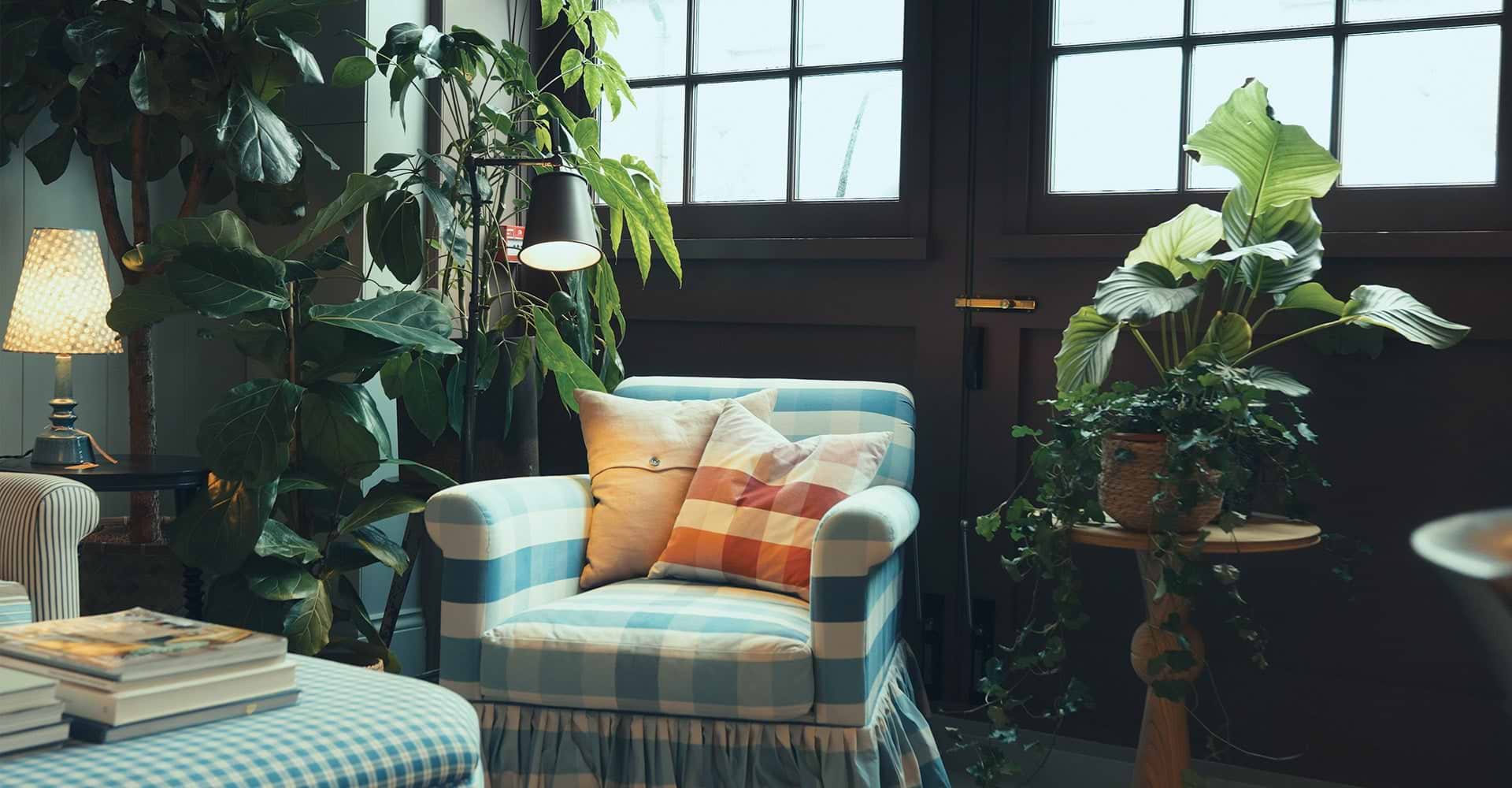 A cozy blue checkered armchair with colorful pillows, surrounded by lush plants and soft lighting, creates a tranquil indoor space.