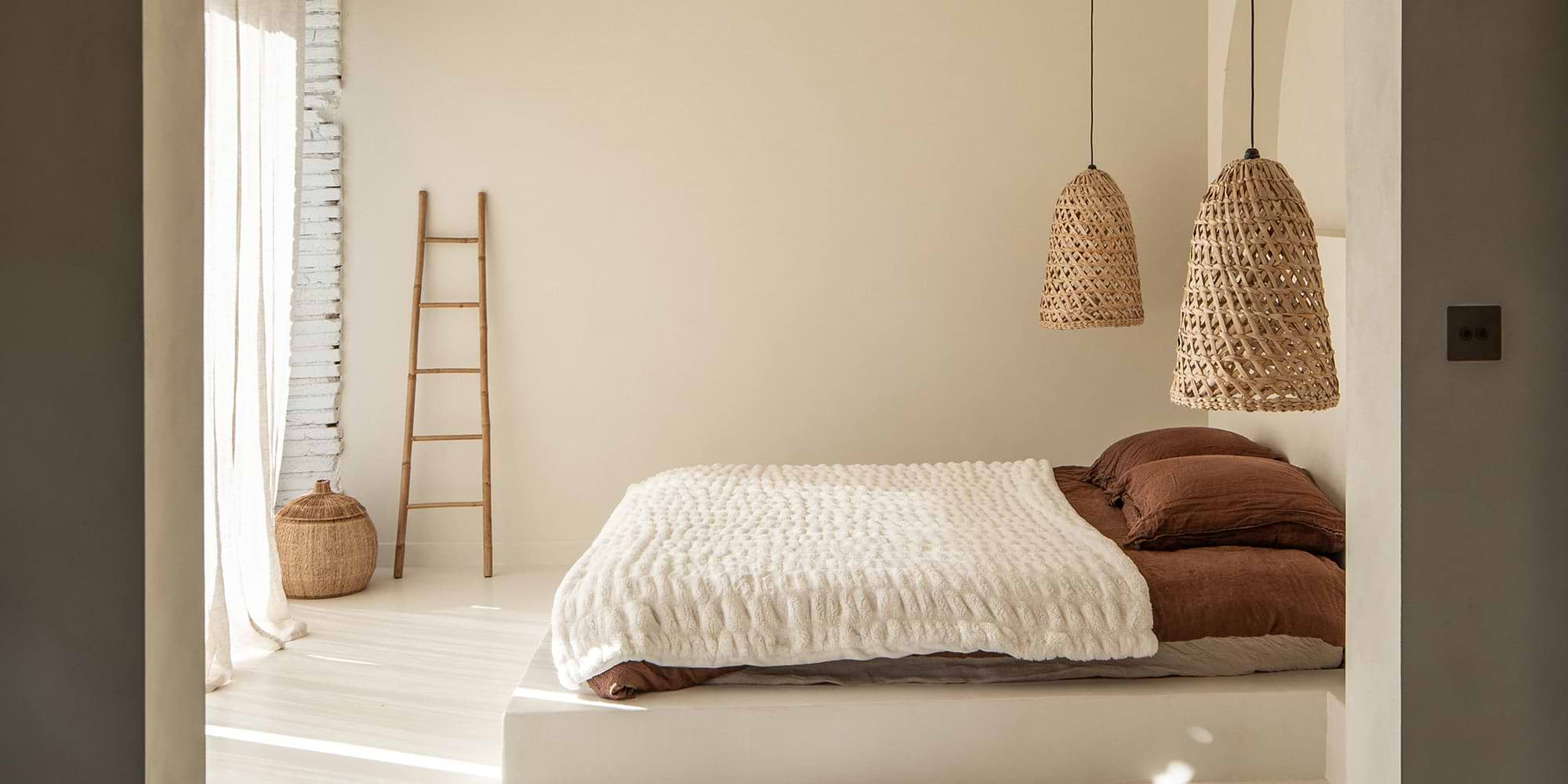 Cozy, neutral-toned bedroom with a textured white blanket, brown pillows, hanging wicker lights, and a wooden ladder against a wall.