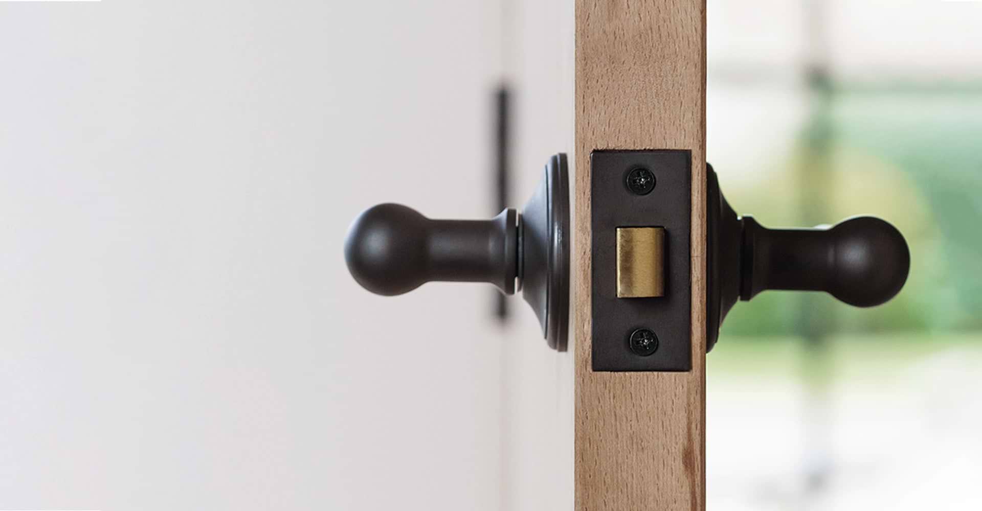 Close-up of a door handle and latch on a wooden door, featuring a round black knob and a brass plate. Soft background blur.