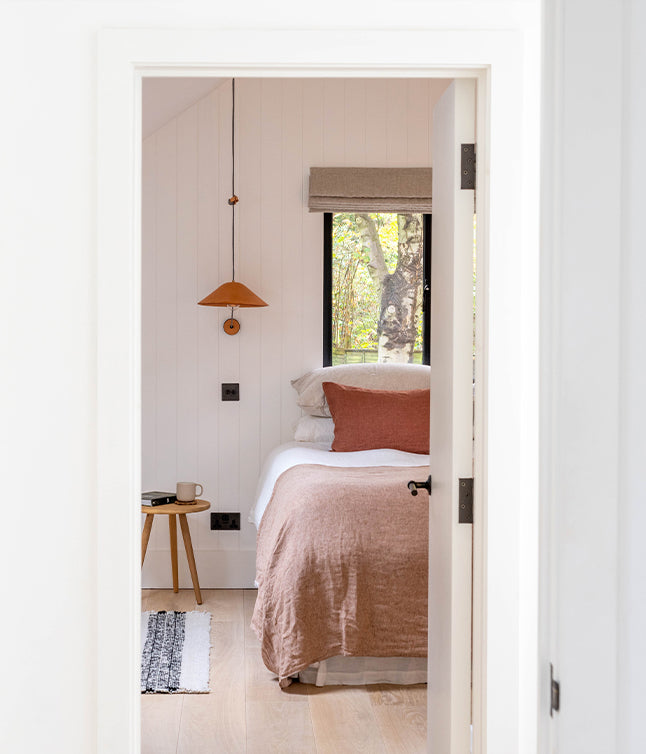 A cozy bedroom view with a warm color palette, featuring a bed, pendant lamp, side table with a cup, and natural light from a window.