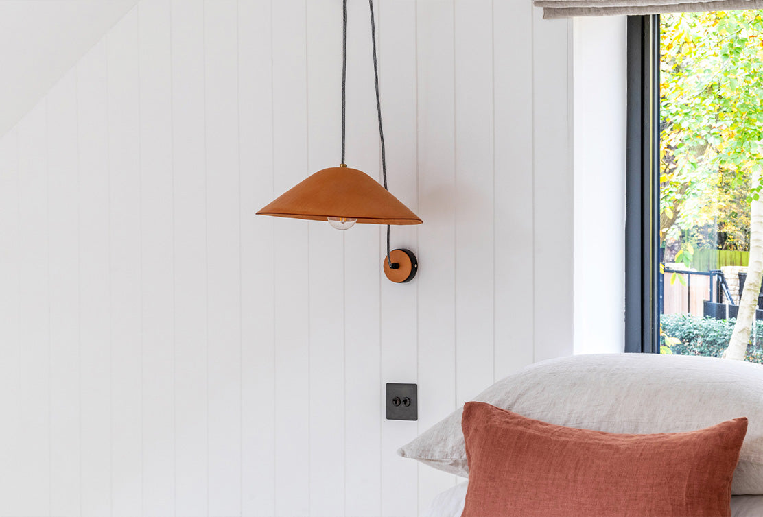 A modern room featuring a brown pendant lamp and a round wall sconce on a white panel wall, with soft pillows and a window view.