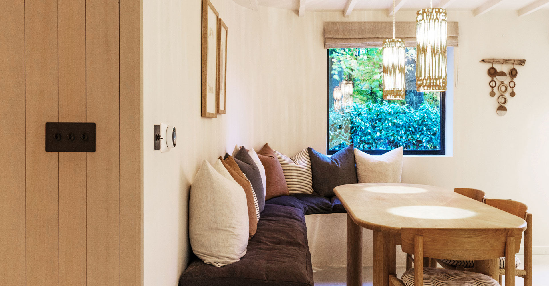 Cozy dining area featuring a plush banquette, natural wood table, soft lighting, and a view of greenery outside the window.