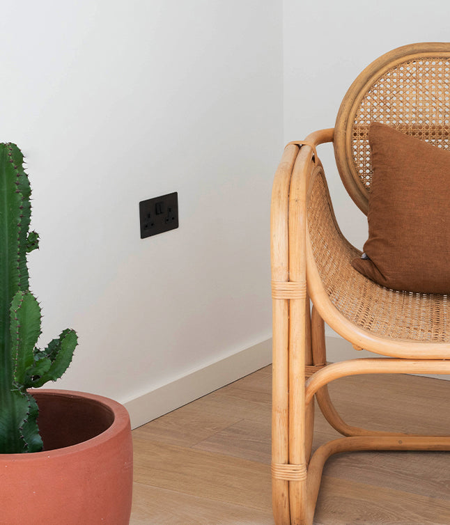 A stylish rattan chair with a brown cushion beside a potted cactus and a bronze electrical outlet against a white wall.