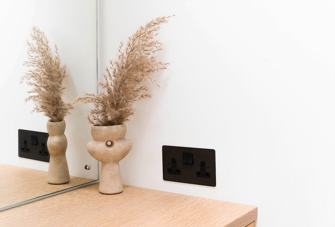 A sculptural beige vase filled with dried pampas grass reflects in a mirror beside a bronze electrical outlet on a light wooden surface.