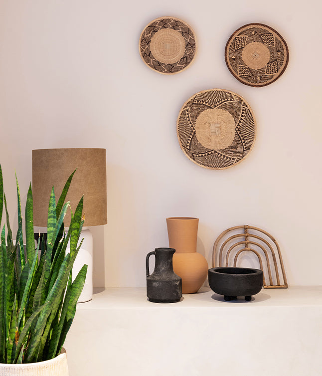 A cozy display featuring woven wall decor, a beige lamp, ceramic vases, a black pitcher, and lush green plants on a shelf.