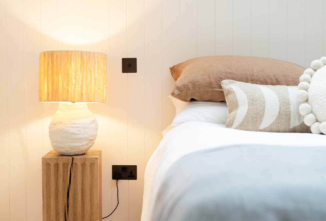 Cozy bedroom corner featuring a textured lamp on a wooden bedside table, plush pillows, and a softly illuminated wall.