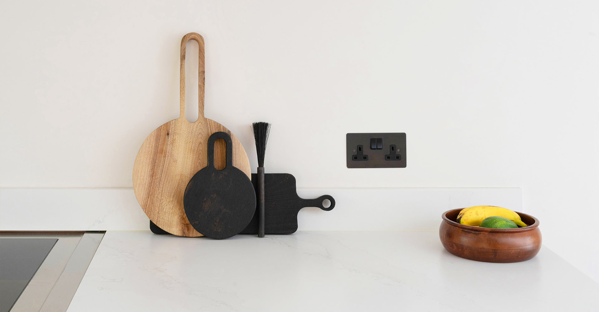 A minimalist kitchen countertop with wooden and black cutting boards, a brush, a power outlet, and a bowl of bananas and limes.