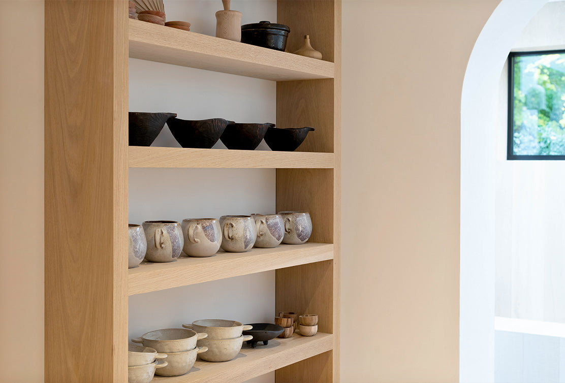 A wooden shelf displays an array of artisanal pottery, including bowls and mugs in various earthy tones and textures against a light wall.