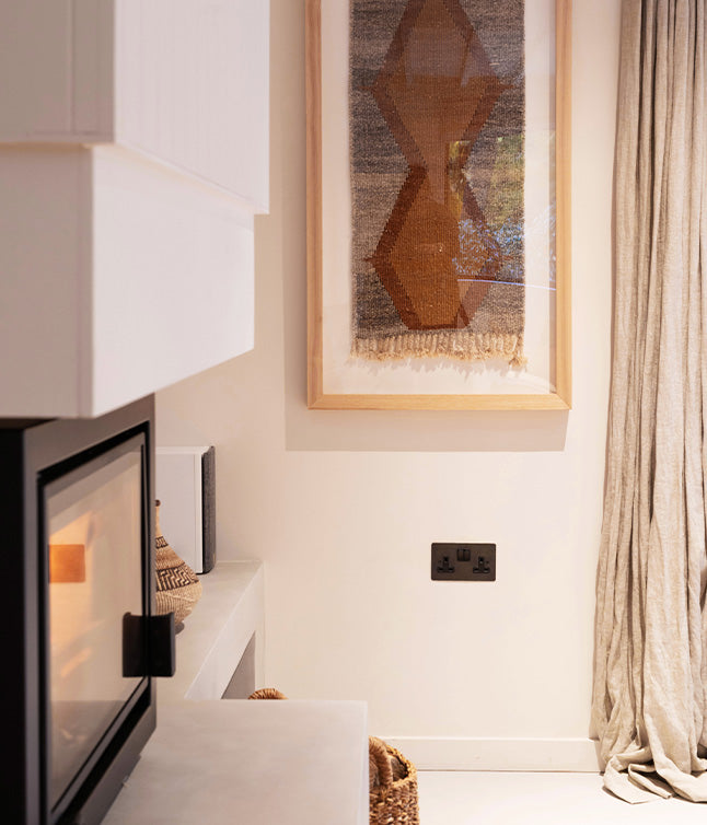 Cozy interior featuring a modern fireplace, textured wall art, soft curtains, and woven baskets on a minimalist white mantel.