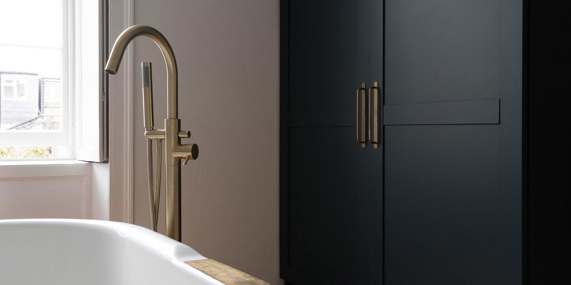 Modern bathroom featuring a sleek gold faucet beside a white bathtub and dark cabinetry against a neutral wall. Natural light filters in.
