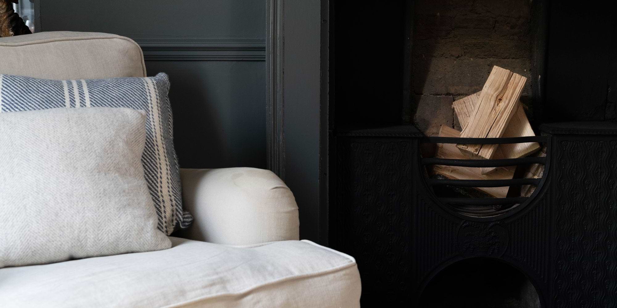 Cozy living room corner featuring a light sofa with decorative pillows beside a black fireplace stacked with firewood.
