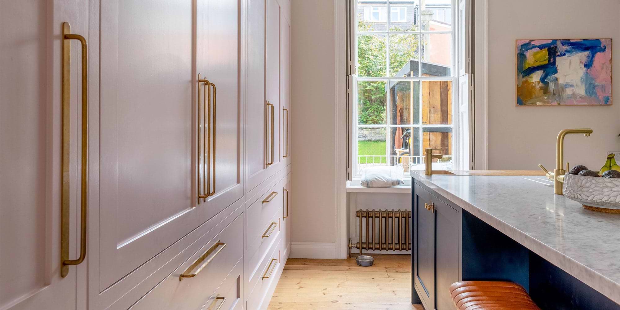 A modern kitchen featuring pink cabinetry, a marble countertop, brass fixtures, and a window overlooking a green garden.