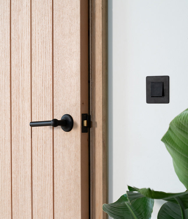 An oak door with a bronze handle, partially open, next to a bronze light switch and a green plant in a modern interior.