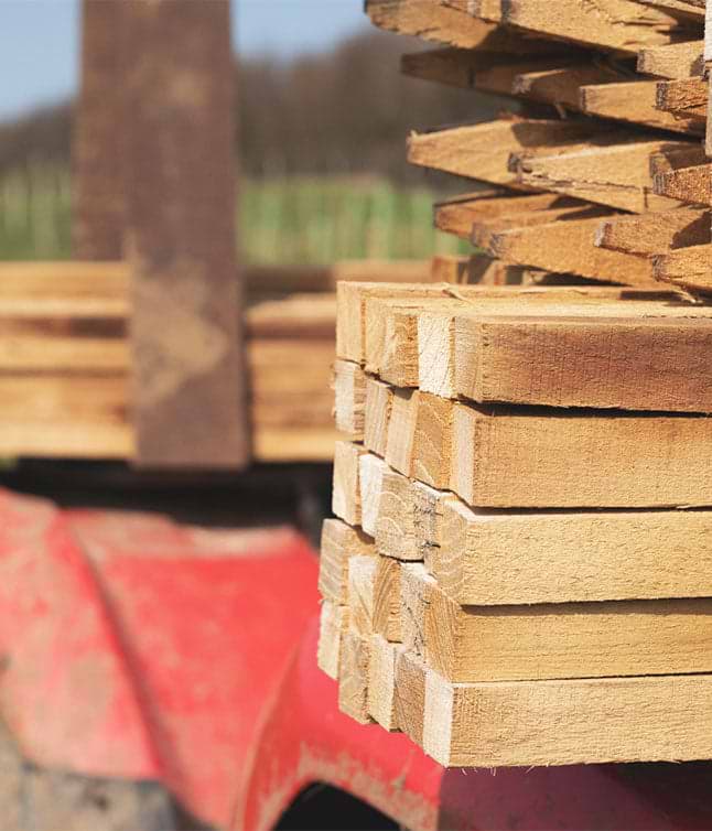 Close-up of stacked wooden planks on a red tractor, with blurred background of green fields and a sunny sky.