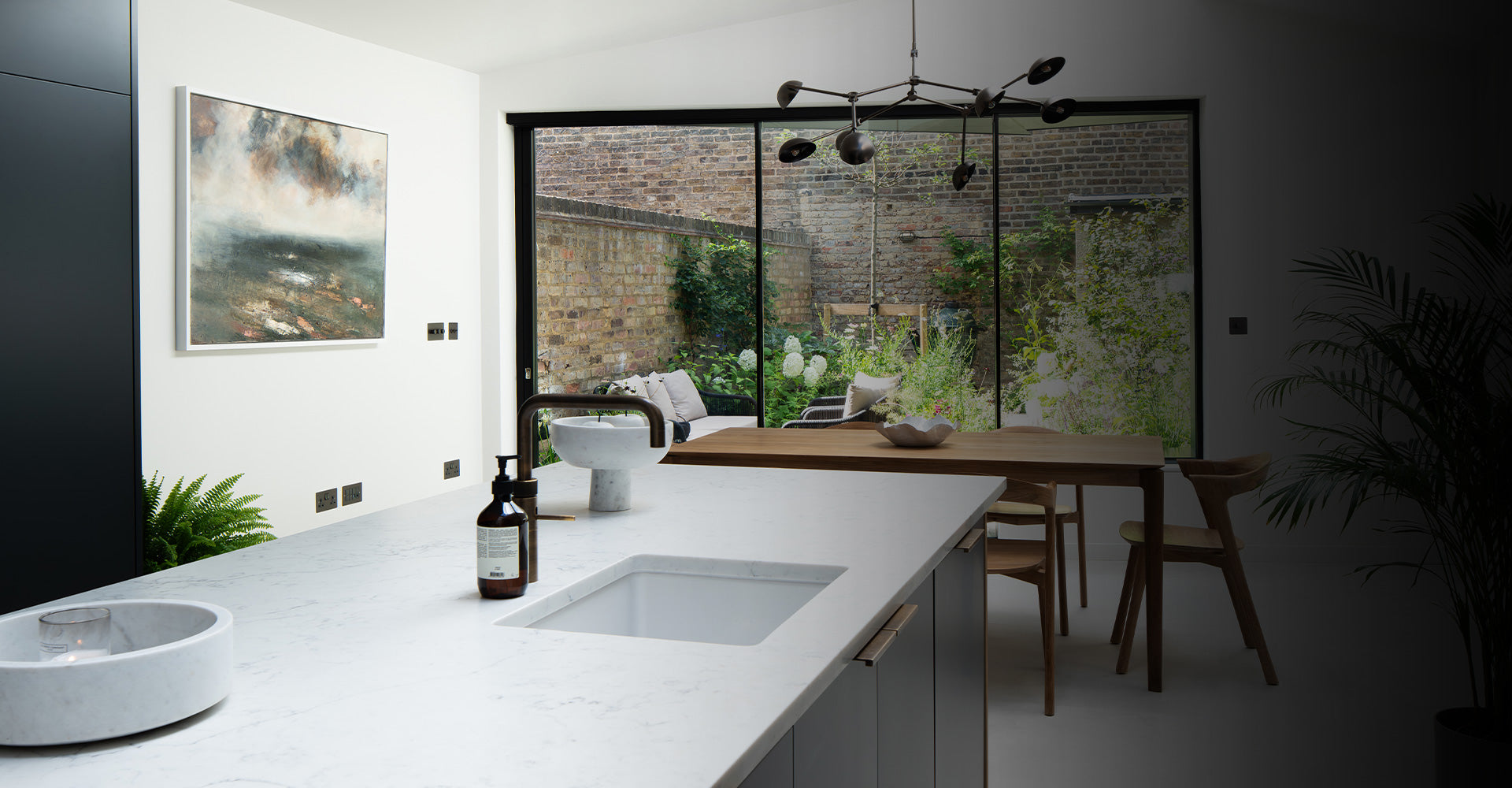 A sleek modern kitchen with a marble countertop, a unique sink, greenery visible through large glass doors, and an artistic landscape painting.