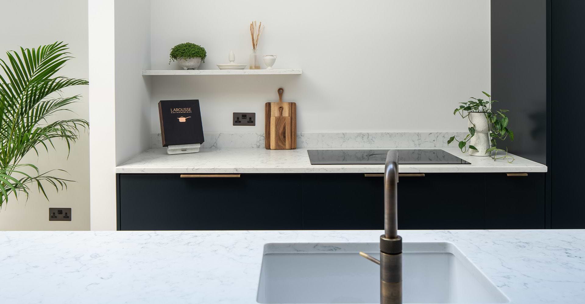Modern kitchen featuring sleek black cabinetry, marble countertops, a cooking surface, wooden cutting board, and decorative plants.