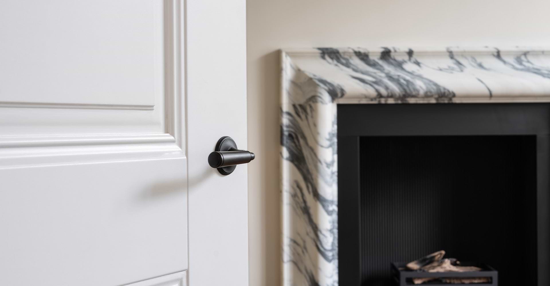 Close-up of a modern black door handle on a white door, with a stylish marble fireplace in the background.