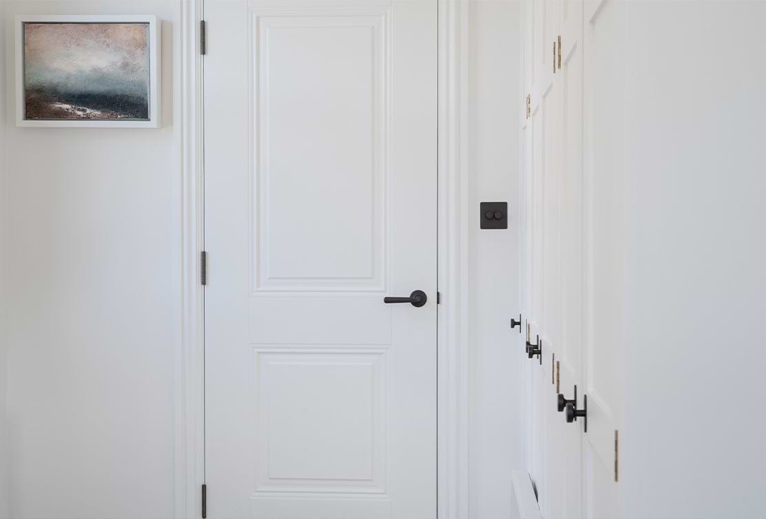 A white door with black handle, adjacent to a light-coloured wall with a small abstract painting and wall switches.