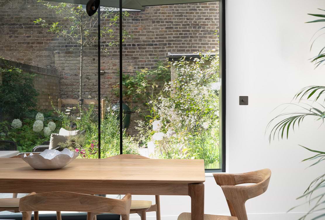A wooden dining table and chair with a silver bowl, overlooking a lush garden through large glass doors.