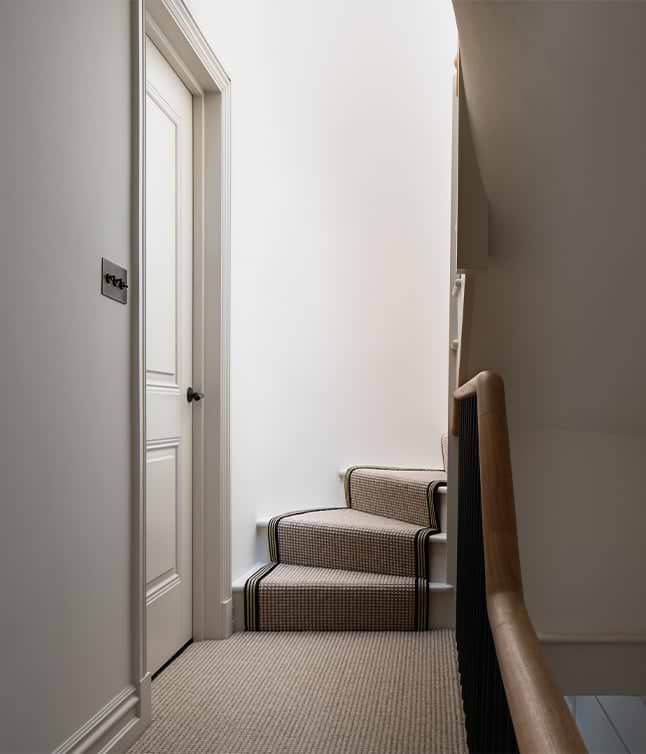 A softly lit staircase with patterned carpet leads to a closed door, featuring a textured railing and minimalist decor.