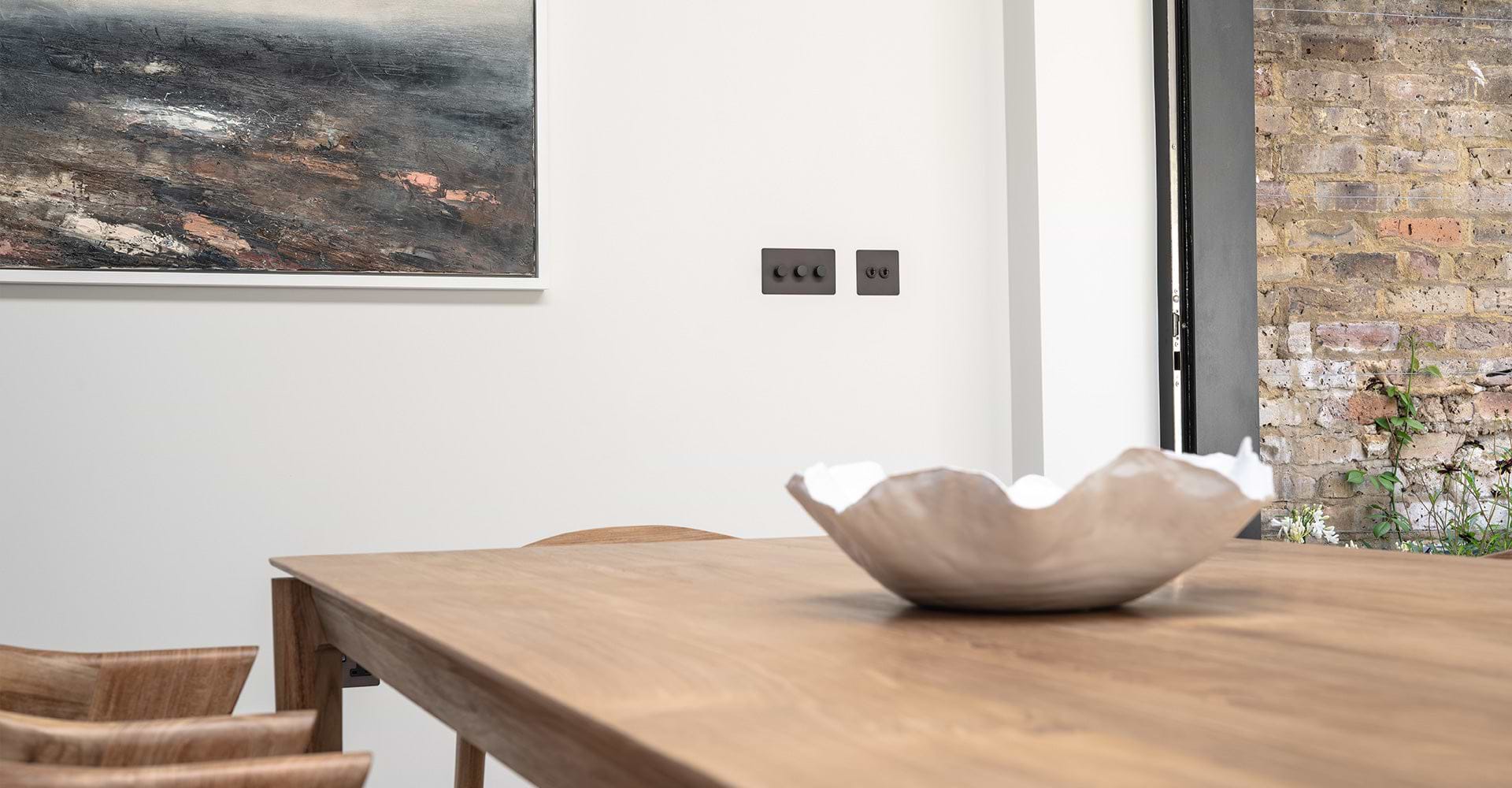 A modern dining area features a wooden table with a decorative bowl and an abstract painting hanging on the wall, alongside light switches.
