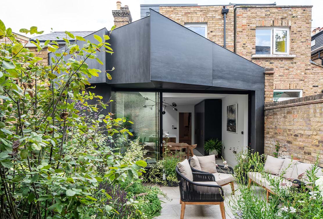 Modern black extension of a home, featuring glass walls, a cozy patio with seating, and lush green foliage surrounding the area.