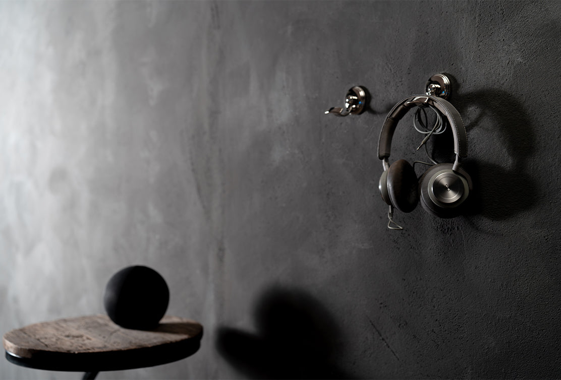 A set of gray headphones hanging on a sleek metal hook against a textured, dark gray wall, with a small black sphere nearby.