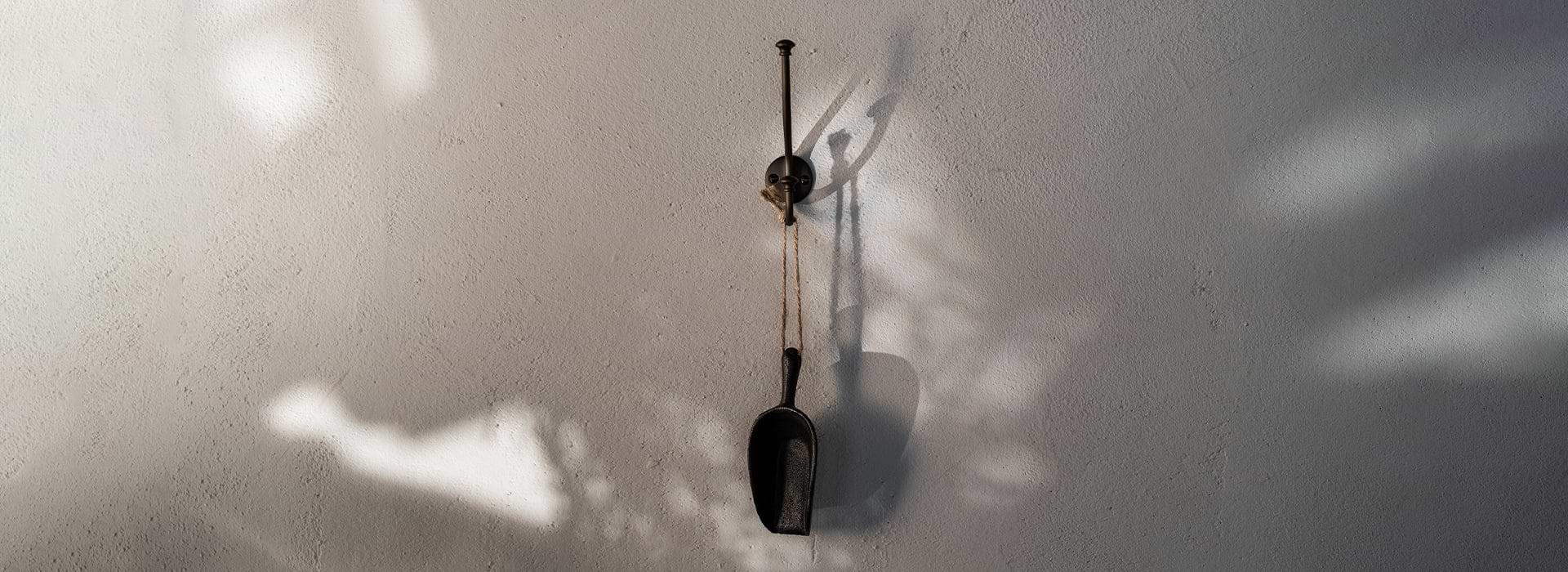 An iron scoop hangs from a black hook on a textured white wall, casting subtle shadows from nearby light.
