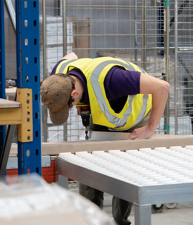 A worker in a yellow safety vest uses a drill to install components on a workbench in a warehouse setting.