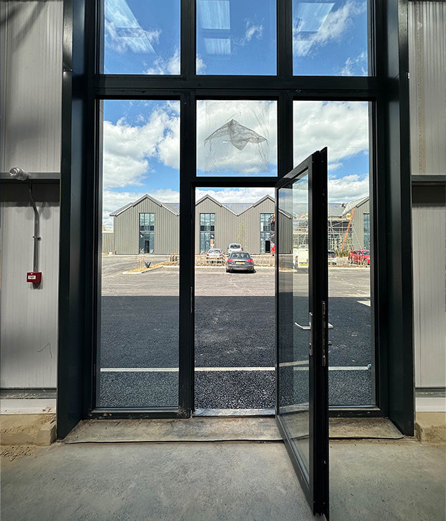 View through an open glass door, revealing modern gray buildings and a blue sky with clouds. A parked car is visible outside.