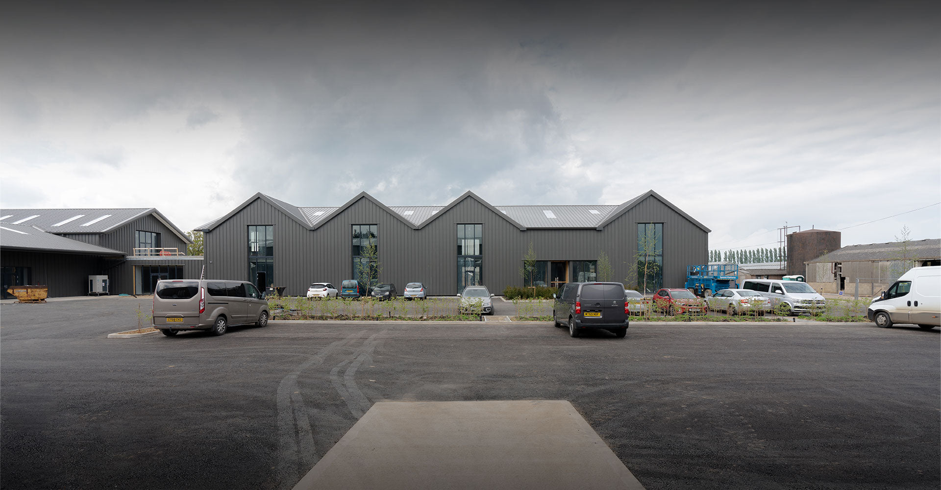 Modern industrial building under construction, featuring multiple vehicles parked outside and green landscaping in front.