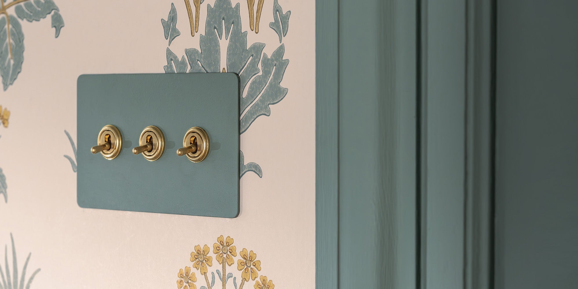 Three decorative light switches with gold fixtures on a teal switch plate, against a patterned wallpaper background.