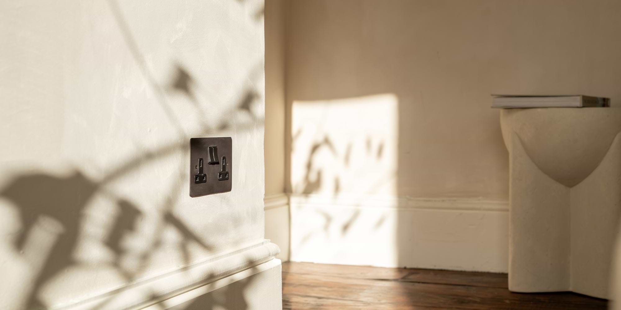 A modern black electrical outlet casts shadows on a light wall, with a minimalist stool and a book adjacent in a softly lit room.