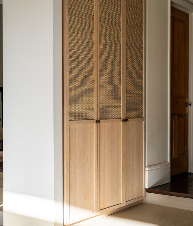 Light wood cabinet with woven rattan doors in a minimalist hallway, illuminated by soft sunlight.