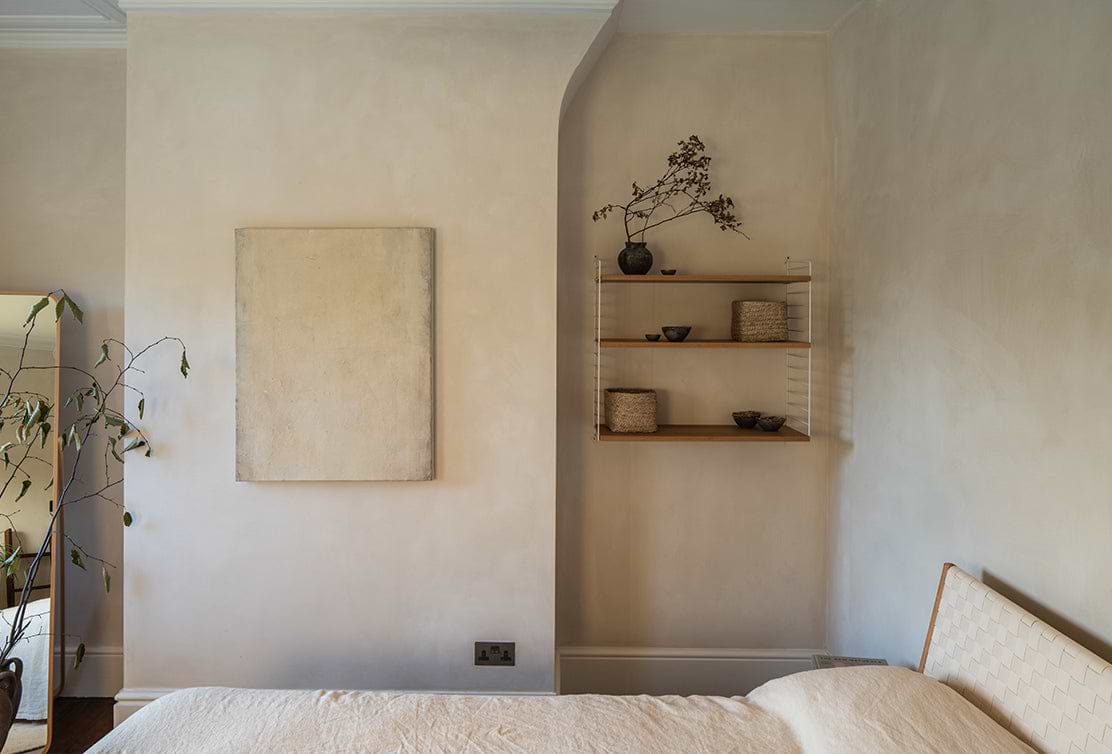 A minimalist bedroom corner featuring a plain wall, abstract artwork, a shelf with decorative items, and a soft-textured bed.