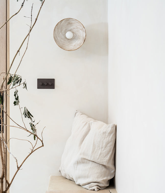 A minimalist corner featuring a textured wall sconce, dark light switch, plush white cushion, and a branch with dried leaves.