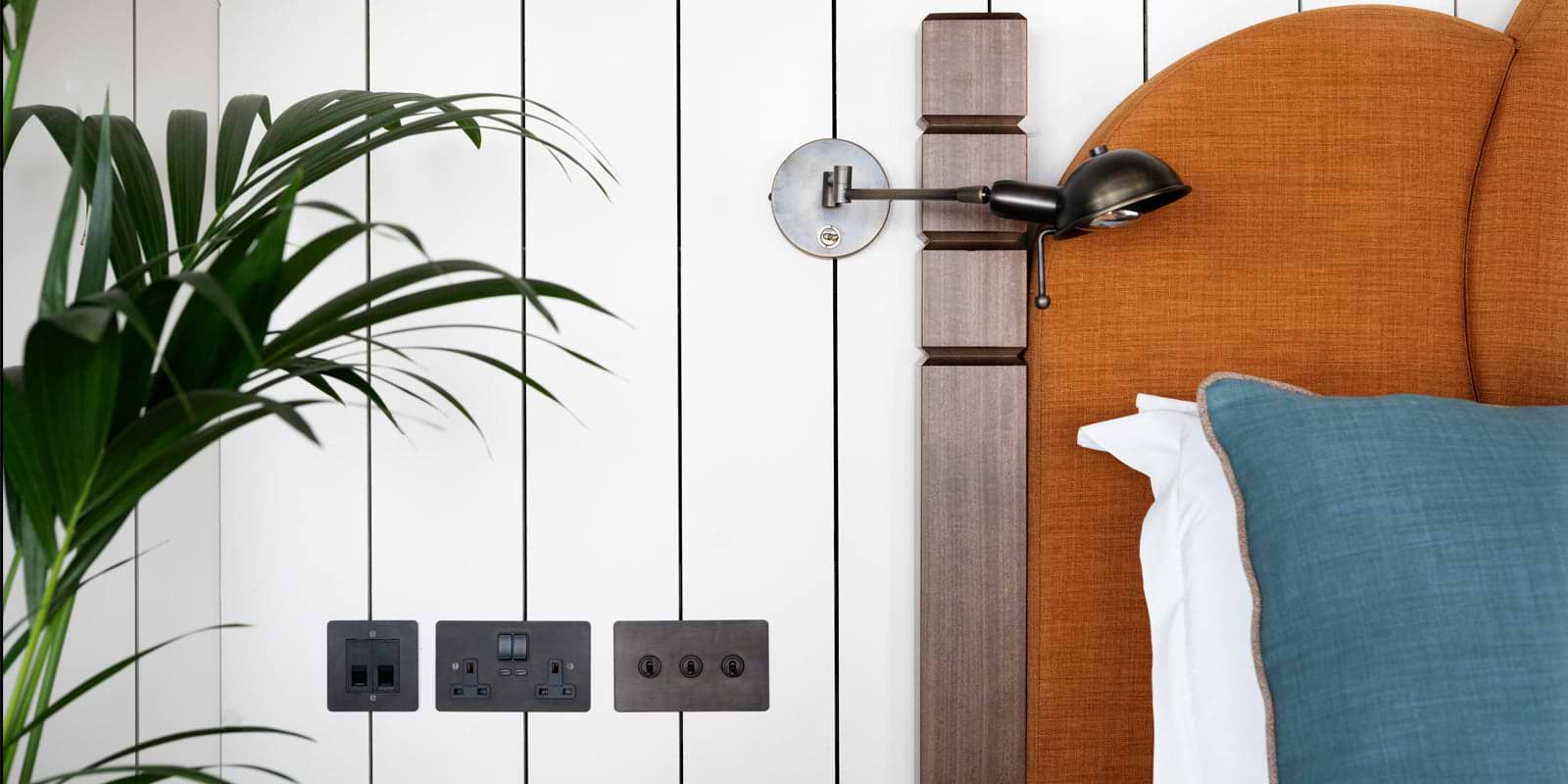 A cozy bedroom corner featuring a stylish orange upholstered headboard, a wall-mounted lamp, and power outlets, framed by lush greenery.