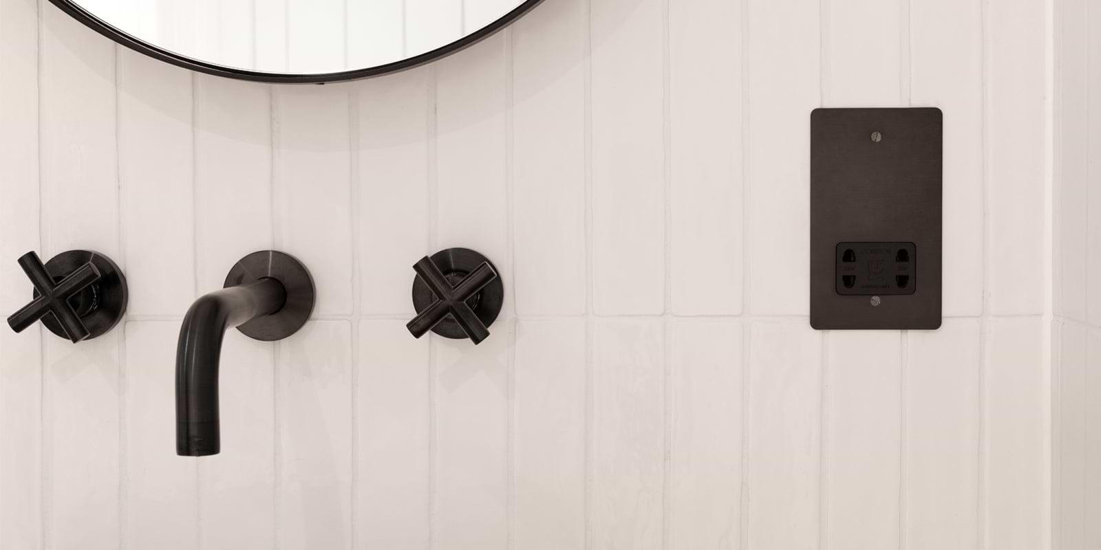 Close-up of a black faucet and wall-mounted controls against a white tiled wall, with a sleek black electrical outlet nearby.