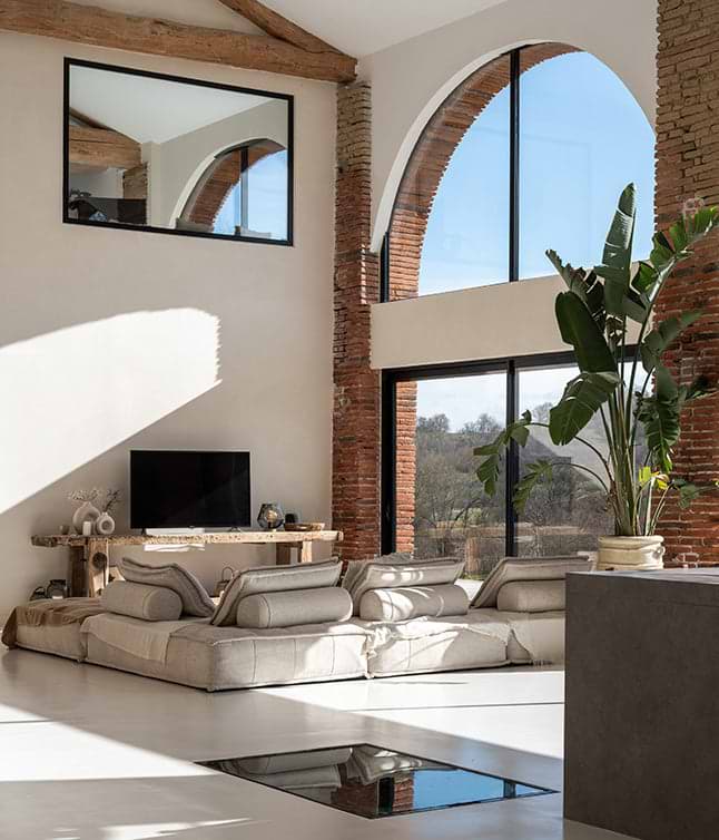 Bright, modern living space featuring large windows, a plush sofa, decorative elements, and greenery against rustic brick walls.